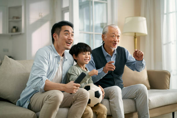 asian son father grandfather watching soccer game on TV together at home asian son father grandfather sitting on couch at home watching live broadcasting of football match on TV together asian kids watching tv stock pictures, royalty-free photos & images