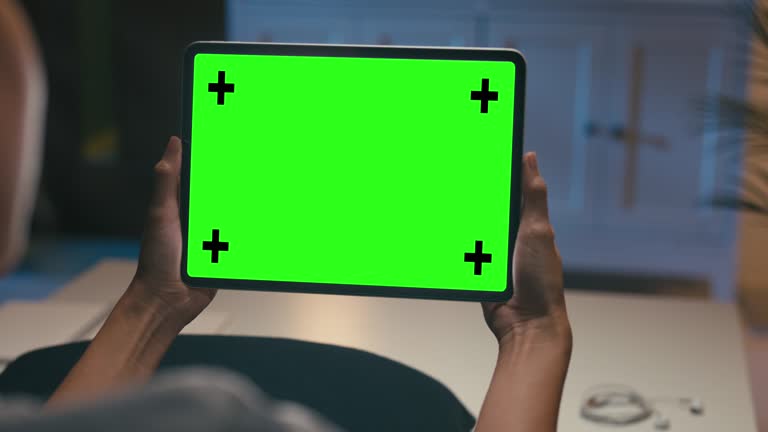Closeup of young Asian woman using digital tablet with mock-up green screen while sitting on couch in living room at home at night. Chroma key technology, Marketing design concept.