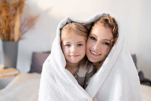 Mom and daughter cutely hugging under a bath towel. Conceptual portrait of a mother and a little cute girl, their wonderful relationship, joint fun and smiles.