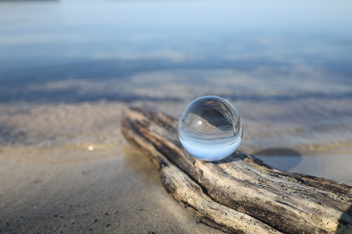 Glass lens ball on driftwood on the beach reflecting the lake and sky