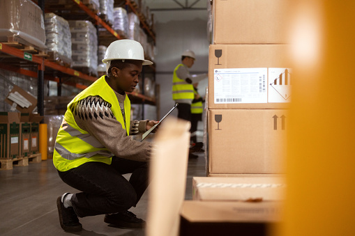 An African-American woman is working in a distribution warehouse taking inventory and moving packages. She is scanning the boxes with a barcode reader.