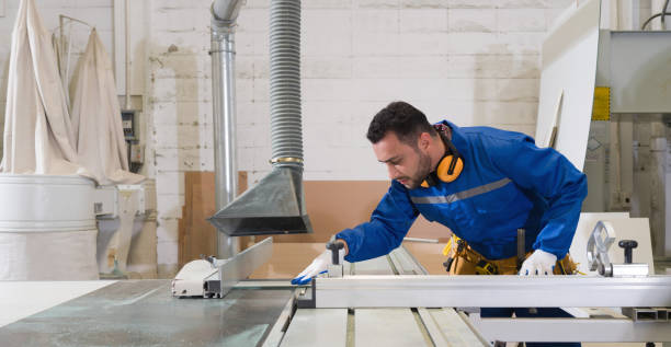 istock Young technician in blue mechanic jumpsuit, ear muff and yellow tool belts inspecting operation of large sliding table saw machine with dust hood. Production line atmosphere in furniture factory. 1450950972