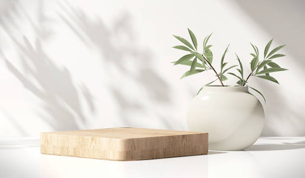 Modern, minimal square wooden podium with white ceramic potted plant on white counter table in dappled sunlight and shadow on white wall stock photo