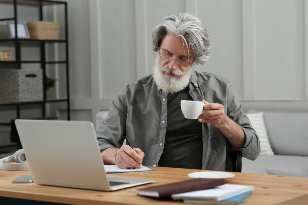 istock Middle aged man with laptop, notebook and cup of drink learning at table indoors 1450962658