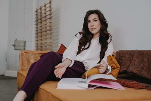 Pensive brunette Spanish woman in white shirt and violet pants sitting on cozy sofa looks aside thoughtfully. Pretty Italian Businesswoman relaxing wt home with opened book. Purposeful people.