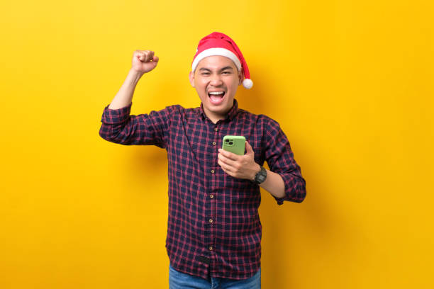 istock Excited young Asian man in Santa hat using smartphone and doing winner gesture over yellow studio background. Happy New Year 2023 celebration merry holiday concept 1450969389