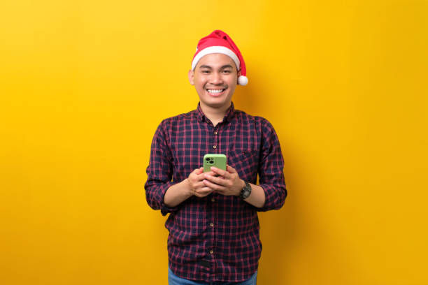 istock Smiling young Asian man in Santa hat holding smartphone, looking at camera over yellow studio background. Happy New Year 2023 celebration merry holiday concept 1450969486