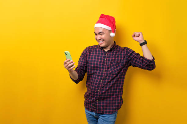 istock Excited young Asian man in Santa hat using smartphone and doing winner gesture over yellow studio background. Happy New Year 2023 celebration merry holiday concept 1450969499