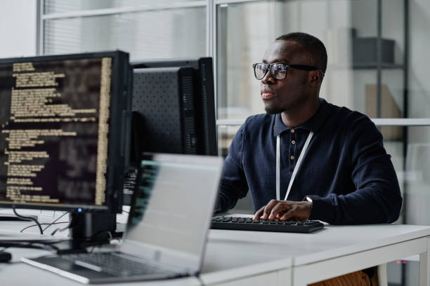 Developer working with new program African American young developer in eyeglasses concentrating on his online work on computer sitting at workplace coding stock pictures, royalty-free photos & images