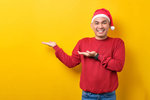 istock Cheerful young Asian man in Santa hat showing empty space on palm over yellow studio background. celebration Christmas holiday and New Year concept 1450969796