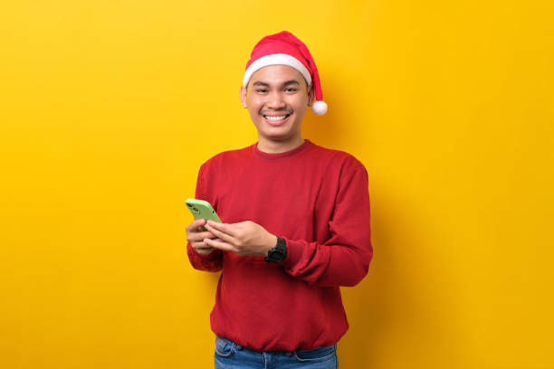 istock Smiling young Asian man in Santa hat holding smartphone, looking at camera over yellow studio background. celebration Christmas holiday and New Year concept 1450970021