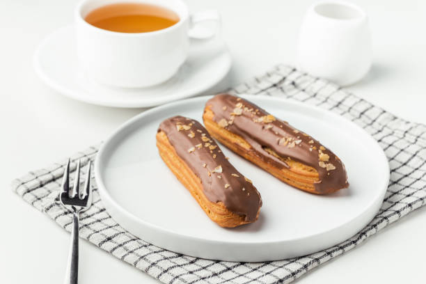 istock Eclairs with custard and chocolate icing on a white plate. Eclairs with cream filling covered with melted milk chocolate. A cup of tea with traditional French pastry in the morning. French breakfast 1450970993