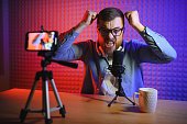 Young man recording or streaming podcast using microphone at his small broadcast studio. Content creator.