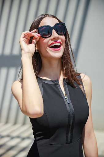 Portrait of beautiful woman wearing black sleeveless top and sunglasses, standing on corporate building terrace and smiling happily on a break