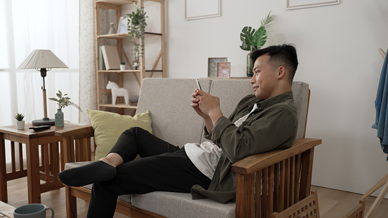 leisure asian young male playing with his mobile phone while reclining on comfortable couch along in the living room at home during daytime.