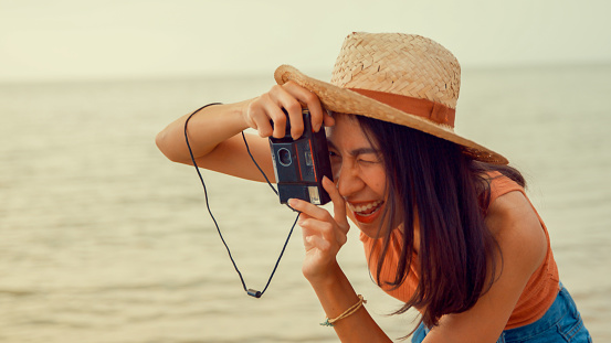 Young Asian woman taking photo picture by retro vintage camera on tropical beach. Holidays vacation concept.
