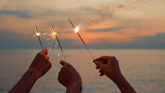 Close up hand of young Asian women celebrating holding sparklers on tropical beach at sunset. Holidays vacation concept.