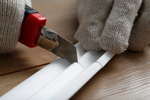 Trabajador cortando molduras de corona de espuma con cuchillo utilitario en mesa de madera, primer plano photo