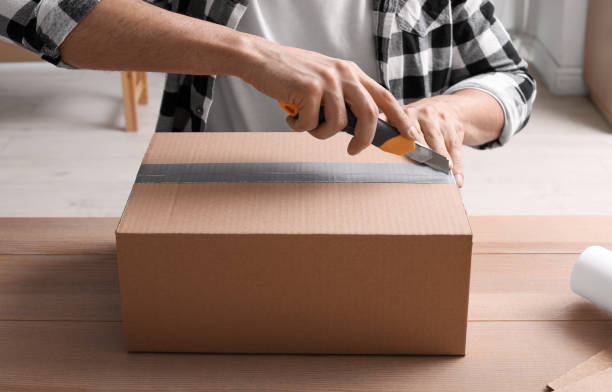 istock Man using utility knife to open parcel at wooden table, closeup 1451003056
