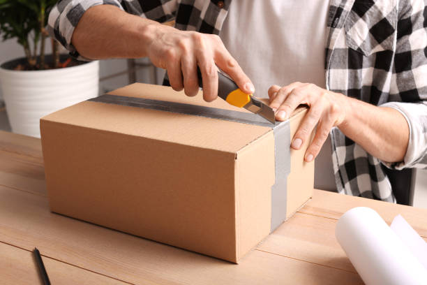 istock Man using utility knife to open parcel at wooden table indoors, closeup 1451004439