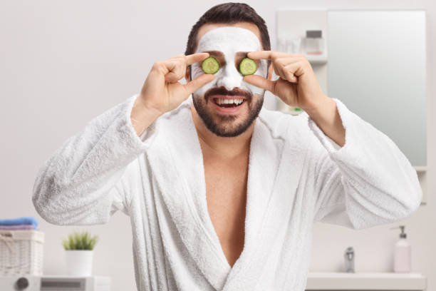 istock Man in a bathrobe with a face mask holding cucumbers on eyes in a bathroom 1451020818