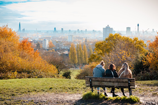London, UK - 28 November, 2022: rear view of three women sitting together on a bench outdoors in the lush and leafy surroundings of Hampstead Heath in London, UK. Beyond them is the city skyline and modern towers and skyscrapers of the London financial district.