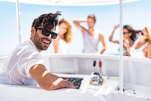 Young happy man driving a boat in summer day while his friends are in the background.