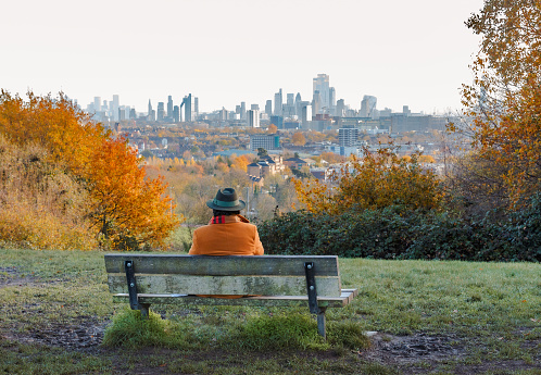 London, UK - 28 November, 2022: rear view of a senior man sitting on a bench outdoors in the lush and leafy surroundings of Hampstead Heath in London, UK. Beyond him is the city skyline and modern towers and skyscrapers of the London financial district.