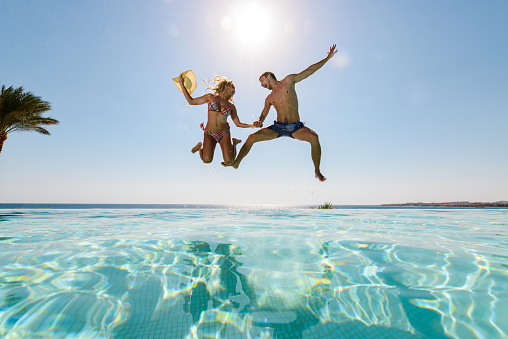 Low angle view of cheerful couple having fun while holding hands and jumping in the swimming pool. Copy space.
