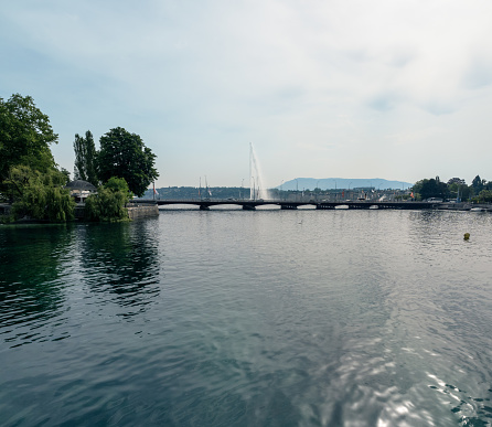 View from the Ponte des Bergues bridge over the waters of the Lake Geneva to the bridge Pont du Mont-Blanc, in the background the attraction Jet d'Eau, the famous fountain in Geneva. On the left part of the island Île Rousseau. 05/22/2022 - Pont des Bergues, 1204 Geneva, Canton of Geneva, Switzerland