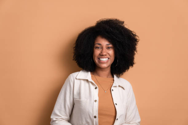 black woman smiling , portrait of real people. happy black young woman smiling and standing wearing white jacket in beige background. portrait, real people concept. natural hair stock pictures, royalty-free photos & images