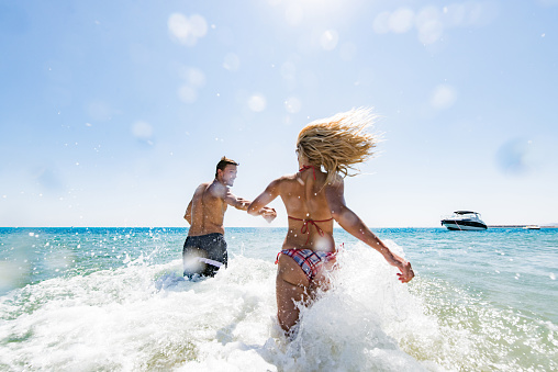 Back view of happy couple having fun while holding hands and running through sea in summer day. Copy space.