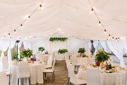 Wedding setup. The wedding arch is decorated leaves and a garland with lights in tent. Luxury elegant tables setting dinner and chairs.  Banquet hall with decor flowers, herbs, citrus fruits lemons.