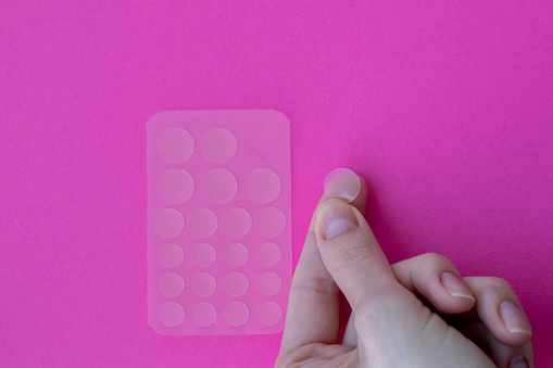Acne patches on a pink background. One patch on a woman's hand, ready to stick to problem skin