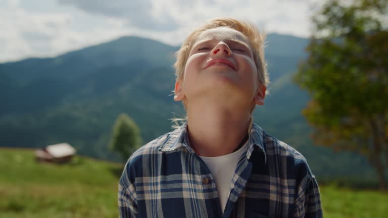 Little boy enjoying sunlight raising head to sky in summer mountains close up.