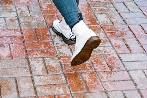 A person wears big white boots at the wet sidewalk tiles of the street. Rain. Wet weather. Bad. Strolling. Sole. Walking. City. Urban