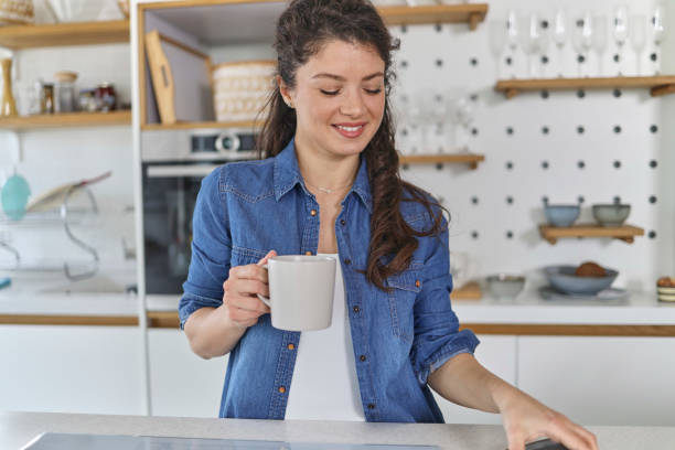 istock Smiling young woman using her mobile phone while drinking a cup of coffee in the kitchen at home 1451067970
