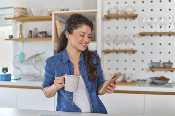 istock Smiling young woman using her mobile phone while drinking a cup of coffee in the kitchen at home 1451068028