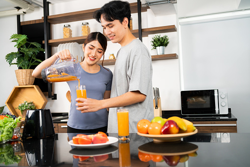 Asian woman pouring smoothies for a man. Happy healthy Asian couple using a blender machine for making healthy vegan smoothies. Couple making vegan smoothies together at home for a healthy lifestyle.