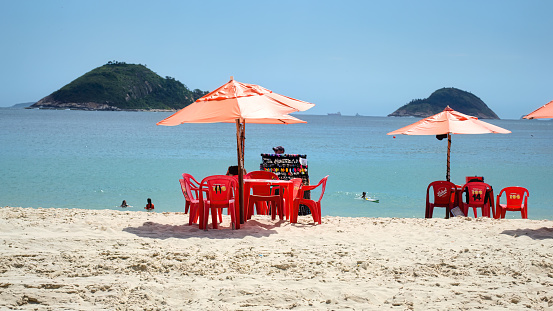 Niteroi, Brazil - December 12, 2022: Red plastic chairs under umbrellas on the beach on a sunny day. Incidental people are in the scene; mountains are in the background.