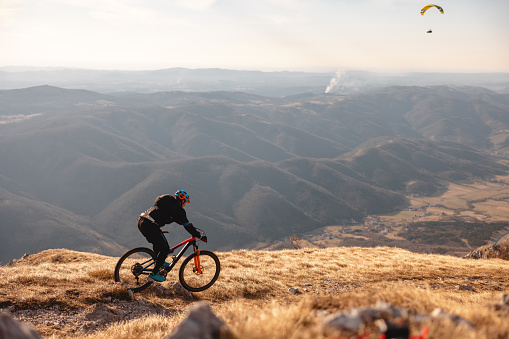 An adult caucasian male athlete riding a mountain bike on top of the hill surrounded by beautiful nature. The athlete has an amazing view of the surrounding hills, mountains and valleys. There is paraglider gliding across the sky above the mountain biker. The male is surrounded by dry grass fields.