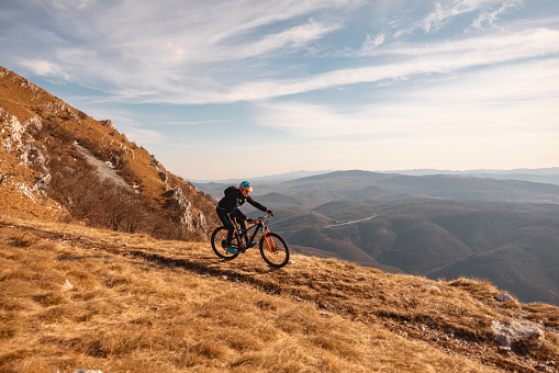An adult caucasian male mountain biker riding downhill. He is located on a beautiful hill with an amazing view. He is wearing a black sports attire and a blue and orange helmet. The sky is blue with some clouds.