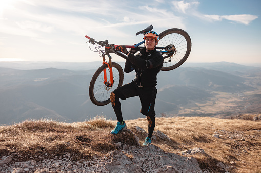 A portrait of a tired adult caucasian male mountain biker standing on top of the hill with an expensive mountain bike on his shoulders. He is looking up as he is resting and standind still. Behind the male there is an amazing view of the beautiful surrounding hills and valleys. The athlete is wearing a colourful helmet that matches the bike and and all black sports attire.