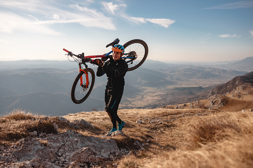 A tired adult caucasian male mountain biker walking while carrying his bike on the shoulder. He is walking to the top of the mountain in order to enjoy the beautiful view as the sun starts to set. The athlete looks content but tired. He is wearing a vibrant helmet and an all black attire. The view behind the male is beautiful. The sun is shining and the sky is blue.