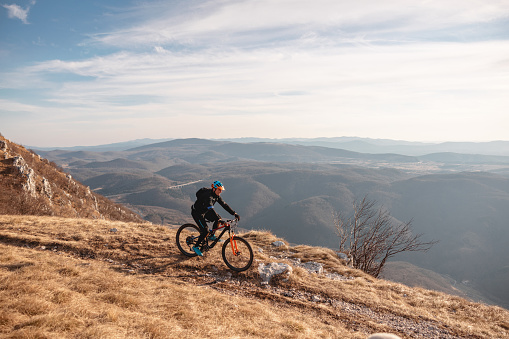 A cool adult caucasian male athlete riding a mountain bike at the top of the hill with an amazing view of the surrounding hills and valleys. The mountain biker is enjoying his ride. The weather conditions at the top of the hill are amazing since the sun is shining and the sky is blue. The athlete is dressed in all black with a vibrant helmet.