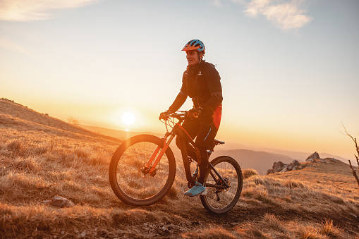A tired adult caucasian male mountain biker slowly biking up the steep bike trail during the sun rise. The man is standing on the bike to bike easier and faster. He is surrounded by beautiful nature and dry grass fields. There is a sunrise and a beautiful view behind the mountain biker. He is wearing a cool vibrant helmet and a black clothing. The sky is clear.