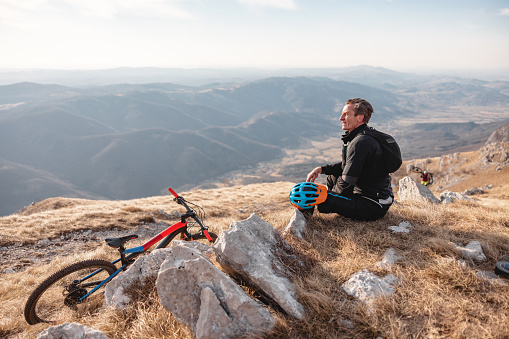 An adult caucasian male athlete sitting on the floor and resting. He has reached the top of the mountain on his mountain biking trip. His expensive mountain bike is leaning agaist a big rock next to the athlete. The athlete is wearing all black. He is admiring the beautiful view of the mountains and valleys. The weather is amazing and sunny.