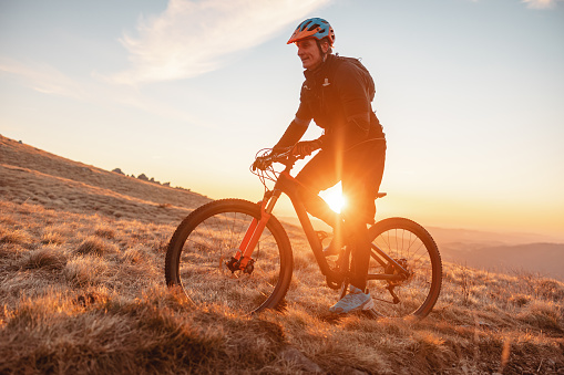 An adult caucasian male athlete riding uphill on a steep bike trail to reach the top of the hill to enjoy the beautiful view of the sunset. The sun is already setting causing a golden hour light that is illuminating the nature surrounding the athlete. The male is wearing a colourful helmet and black clothes.