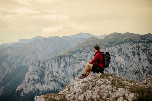 Male hiker with backpack looking at beautiful landscape scenery while sitting on stone at mountain top