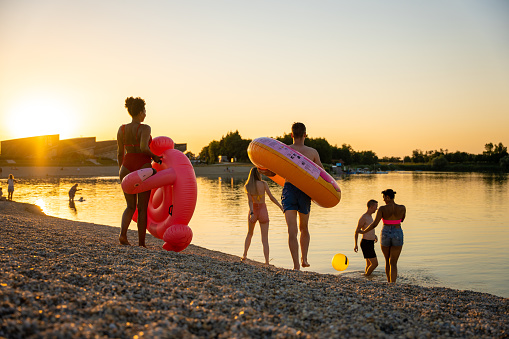 Multiracial young male and females friends with inflatable rings and pool ball enjoying vacations together at lakeshore during sunset